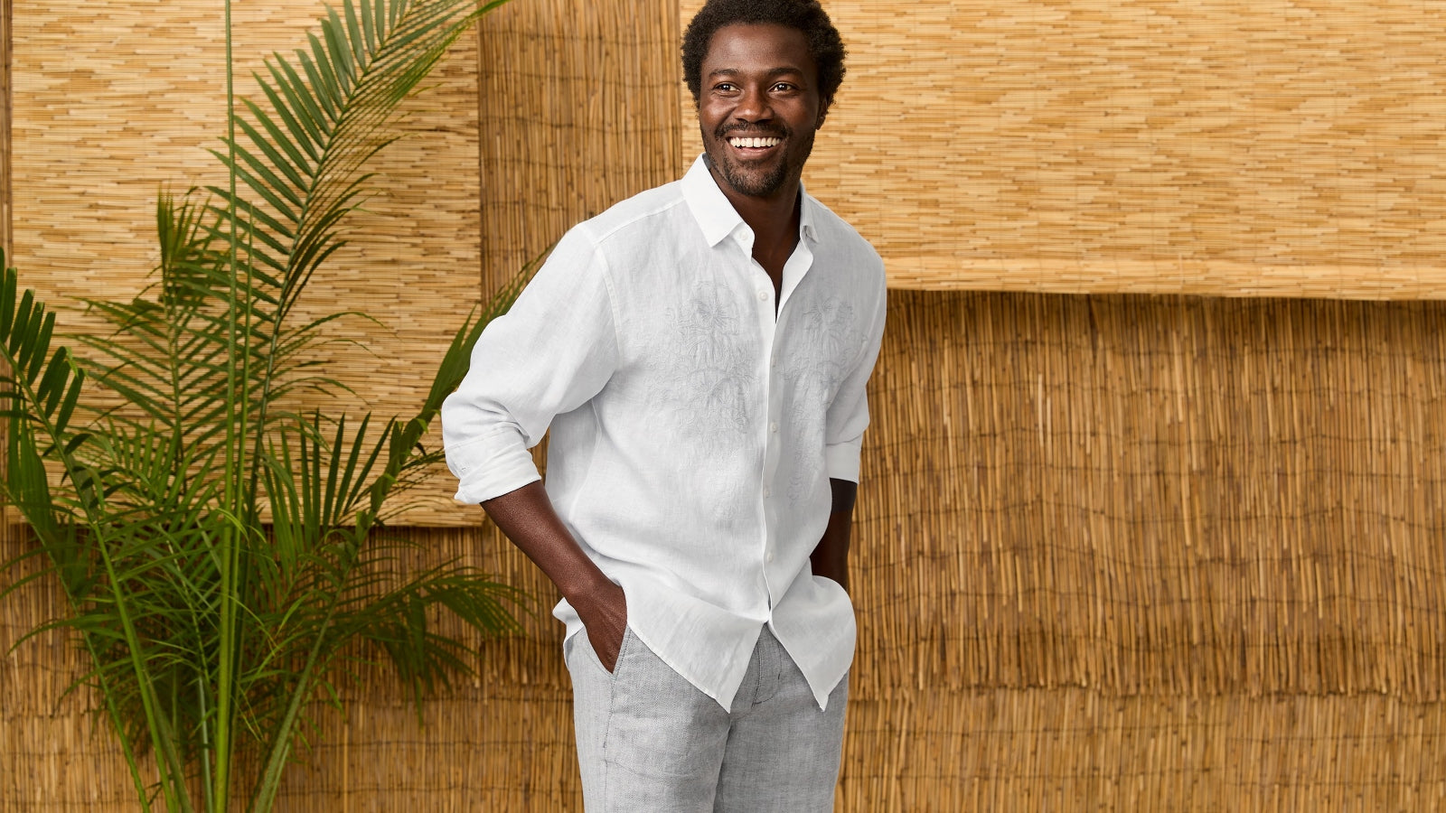 Man wearing a white shirt and pants standing against a wooden wall with plants.