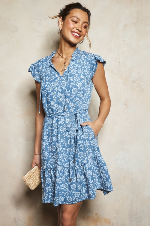 A woman modeling a blue and white floral print mini dress with ruffled cap sleeves, a button-up front, and a tie waist, holding a small woven clutch against a textured beige wall.