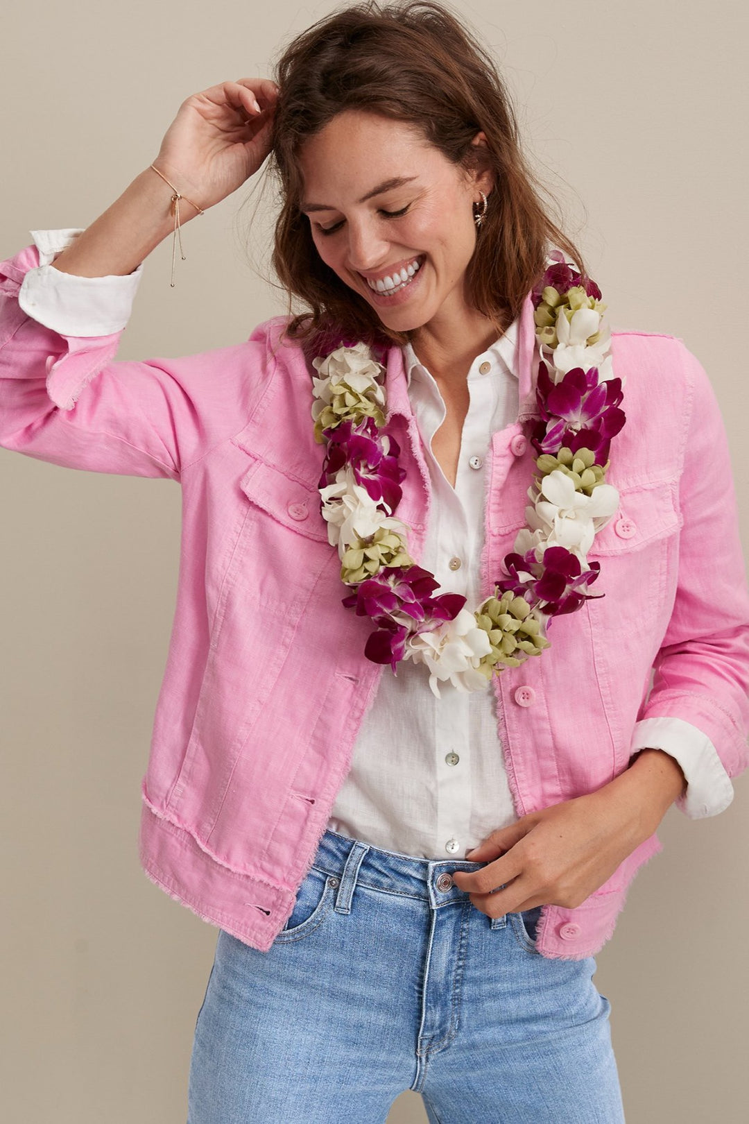 Woman wearing a pink jacket, white shirt, and blue jeans with a floral lei on a beige background