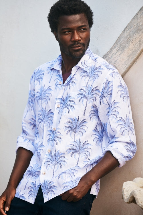 A man wearing a white long-sleeve button-up shirt with a blue palm tree pattern, standing against a textured beige wall.