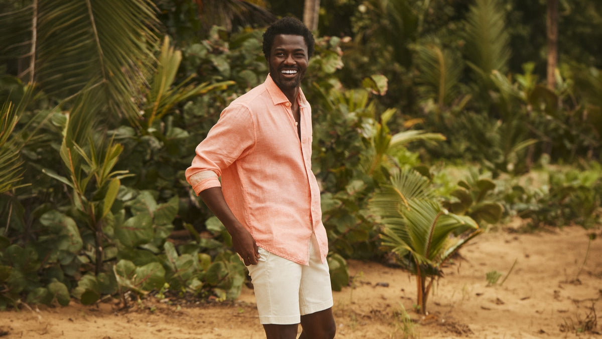 A man standing outdoors in a tropical setting, wearing a light peach-pink long-sleeve button-down shirt and white shorts.