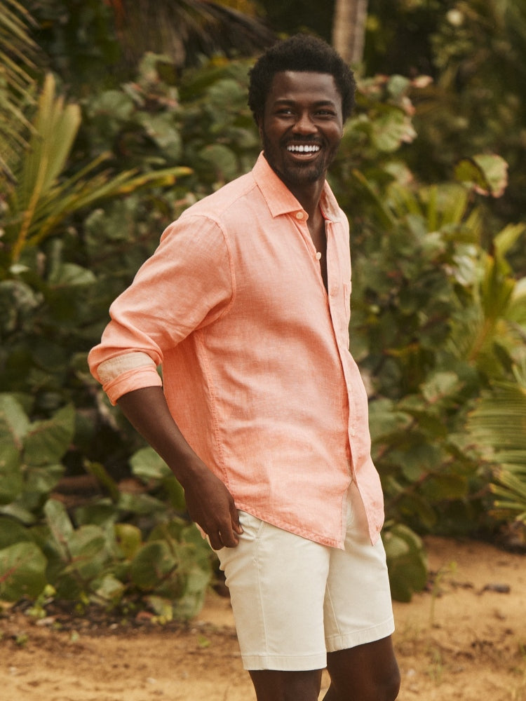 A smiling man wearing a light peach linen button-down shirt and off-white shorts, standing outdoors in front of lush tropical green foliage.