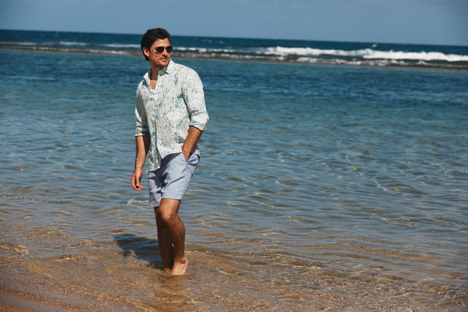 Man walking in shallow water at the beach with sunglasses on