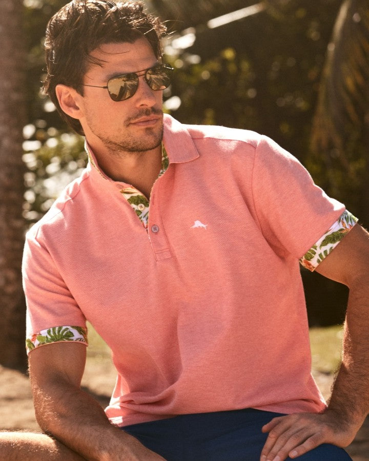 A man wearing a coral pink polo shirt with green tropical leaf-patterned inner collar and sleeve cuffs, styled with dark sunglasses and navy blue pants, seated outdoors in warm sunlight.