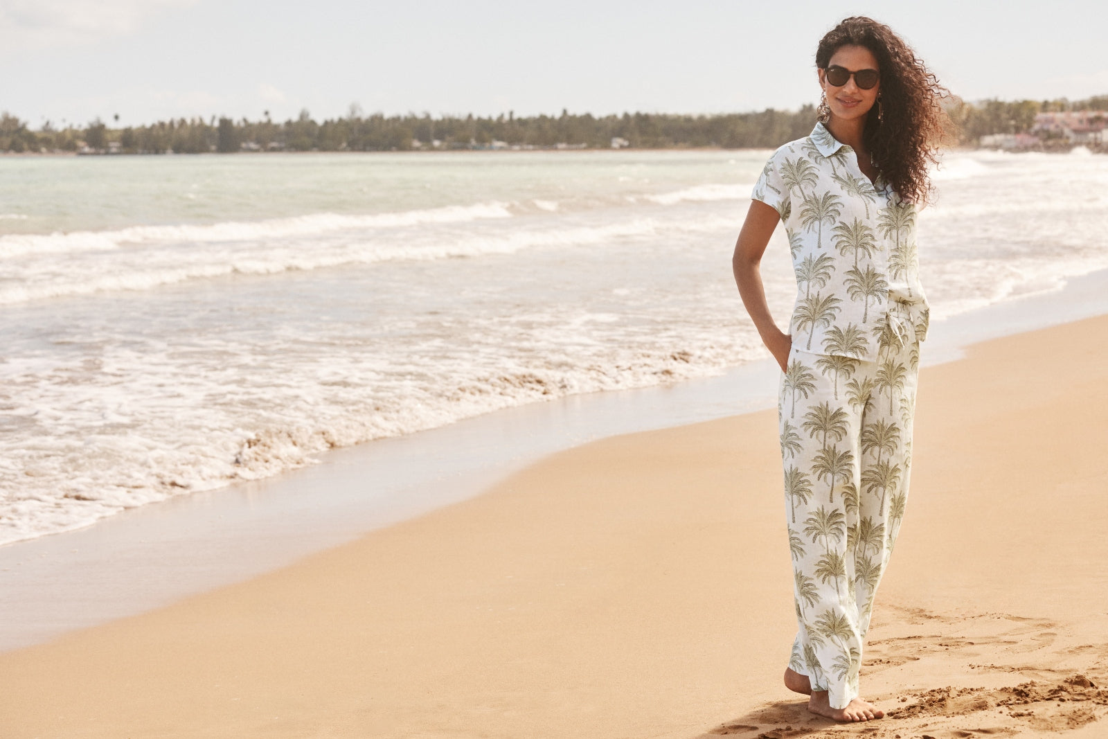 A woman wearing a white short-sleeve collared shirt and matching wide-leg pants with a green palm tree print stands on a sandy beach next to the ocean.